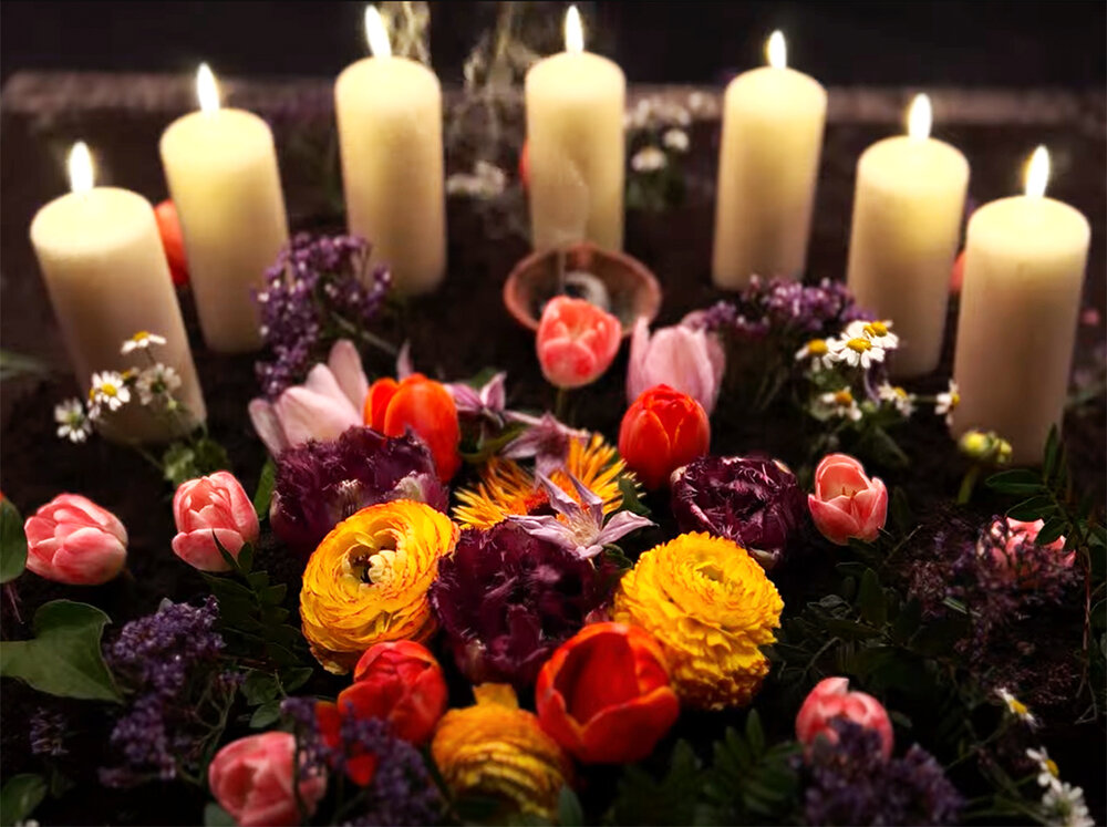 Bouquet of flowers in reddish and yellow colours in the foreground, surrounded by seven burning white candles.